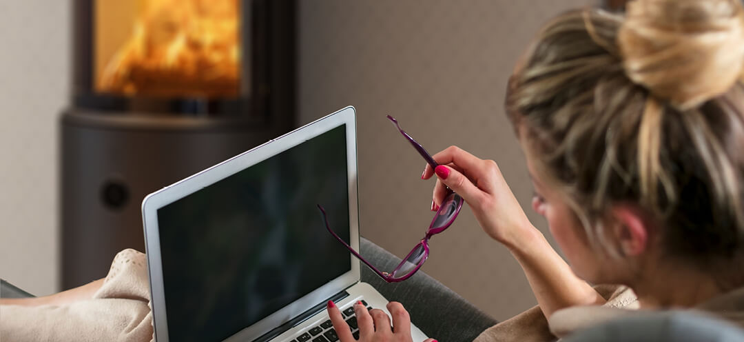 Frau mit hochgestecktem Haar sitzt vor einem schwarzen Holzofen Scan 67, mit einem PC auf dem Schoß und einer Brille in der Hand
