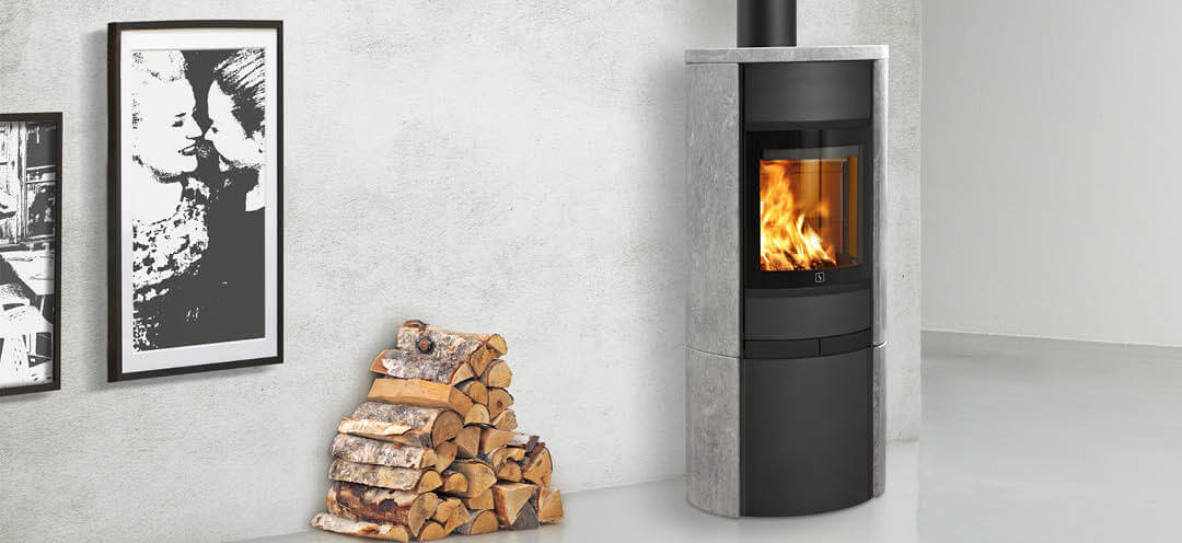 Schwarzer Kaminofen mit Seitenwänden aus Speckstein Scan 68-15 in einem weißen Raum, daneben ein Stapel Holz