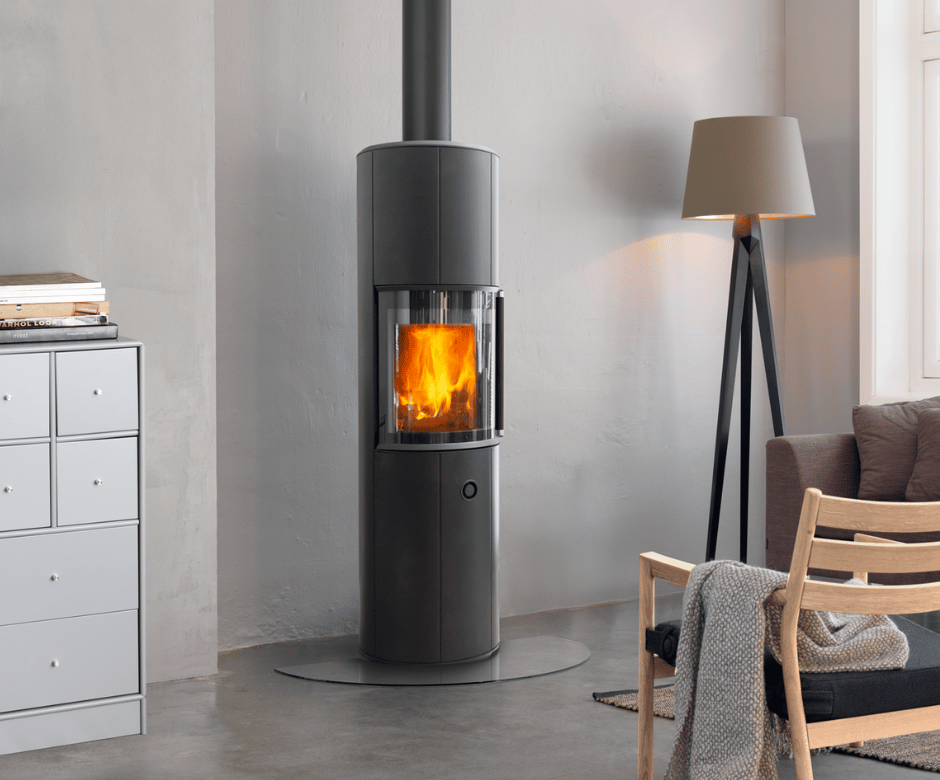 Moderner, zylinderförmiger Scan 67 1600 Zensoric Holzofen mit durch eine große Glastür sichtbaren Flammen, platziert in einem stilvollen skandinavischen Wohnzimmerinterieur mit Lampe, Stuhl und Aufbewahrungsmöbel