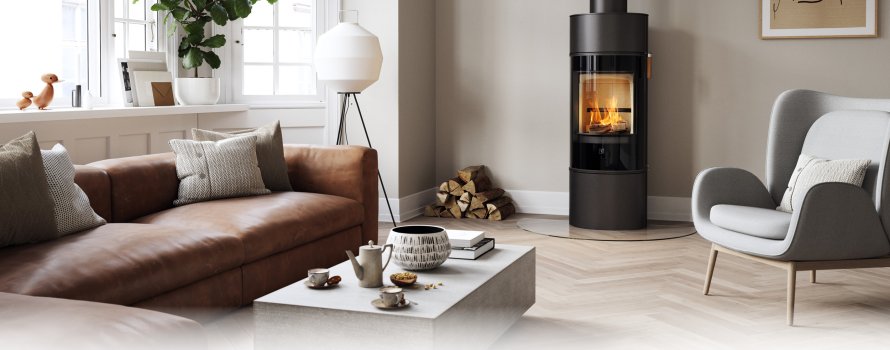 Schwarzer Kaminofen Scan 84 mit schwarzer Glastür In einem modernen nordischen Wohnzimmer mit braunem Sofa und hellgrauem Stuhl