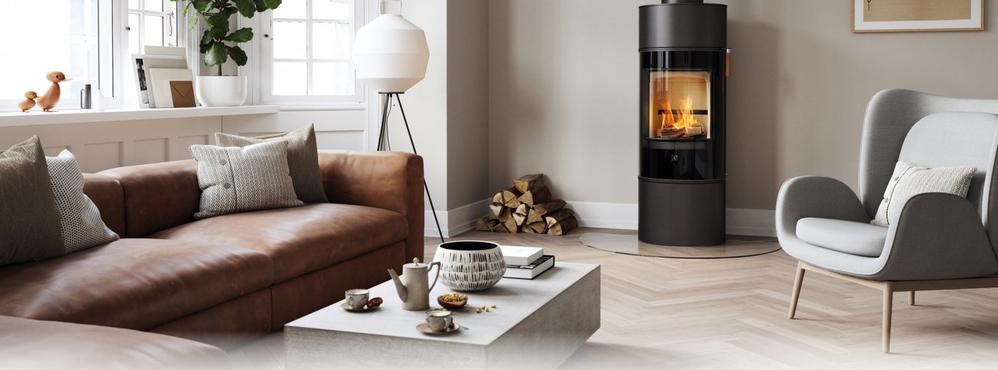 Schwarzer Kaminofen Scan 84 mit schwarzer Glastür In einem modernen nordischen Wohnzimmer mit braunem Sofa und hellgrauem Stuhl